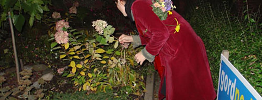A woman in a red coat leaning over picking flowers from a garden in the night. 