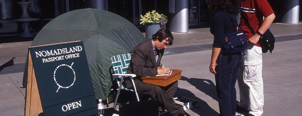 Artist sitting outside tent writing on a paper. Sing to the left says Passports for nomadics. On left two people stand waiting.