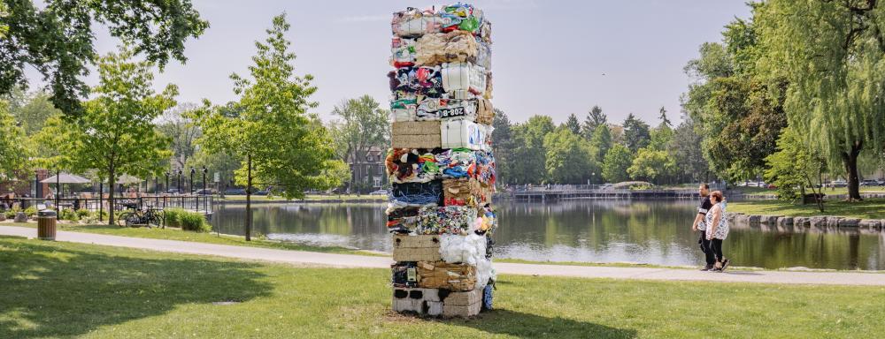 Two people walk on a path beside water, looking toward a tall rectangular structure made out of bales of compacted recyclable waste.