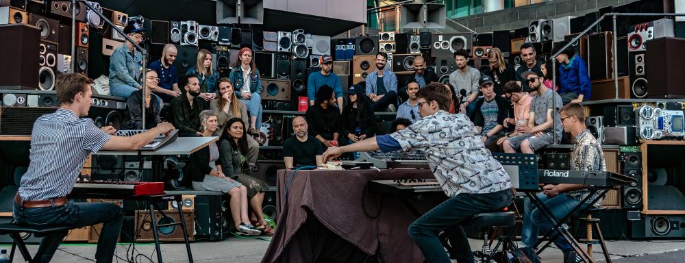 musicians play synths to a crowd seated on arena-style bleachers assembled from 400 loud speakers